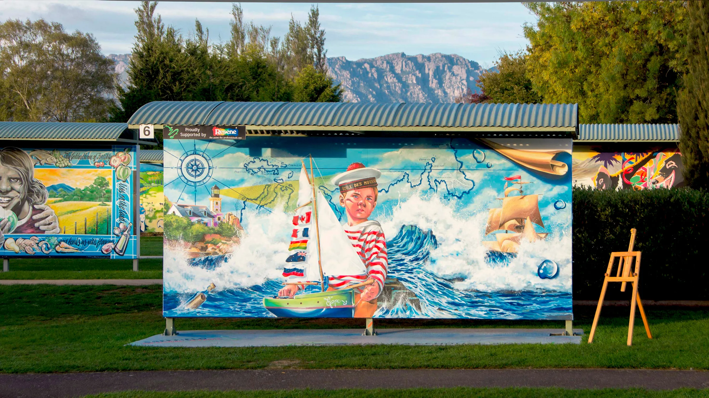 Fresque murale « I'll Journey Soon Around The World » de Gwendal Larher, primée à l'International Mural Fest de Sheffield, en Tasmanie (Australie) : jeune mousse et voilier sur un local de parc entouré de montagnes